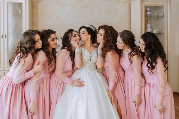 Portrait of the bride with her friends in the room. A brunette bride in a long white dress and her friends in pink dresses are smiling and happy. Young girls.