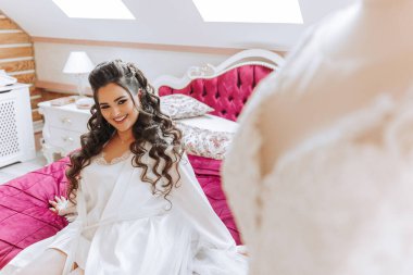 Mutlu bir gelin, bir otel odasında, bir mankenin üzerinde gelinliğiyle lüks düğününe hazırlanıyor. Şık saçlı, makyajlı ve sabahlık giymiş bir kadının portresi..