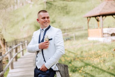 Beyaz ceketli ve siyah pantolonlu damat gülümsüyor ve yaka çiçeğini düzeltiyor. Düğün portresi.