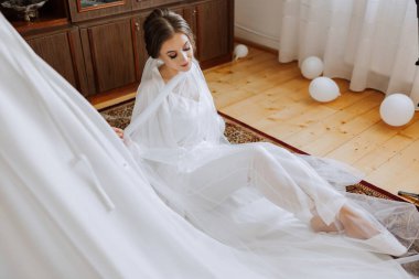 Mutlu bir gelin, bir otel odasında, bir mankenin üzerinde gelinliğiyle lüks düğününe hazırlanıyor. Şık saçlı, makyajlı ve sabahlık giymiş bir kadının portresi..