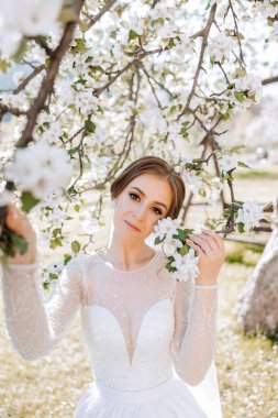 Kızıl saçlı bir gelin çiçek açan bir ağacın arka planında poz veriyor. Uzun kollu muhteşem bir elbise, açık göğüs. Bahar düğünü