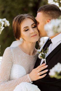 Düğün için. Aşk ve çift düğün için bahçede. Törenin ve bağlılığın kutlanması. Tarihi kaydet. Güven. Gelin ve damat kucaklaşır. Damat gelini bahar çiçekli bir ağacın altında kucaklıyor..