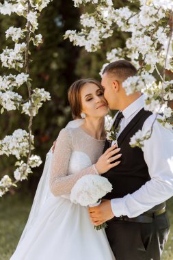 Düğün için. Aşk ve çift düğün için bahçede. Törenin ve bağlılığın kutlanması. Tarihi kaydet. Güven. Gelin ve damat kucaklaşır. Damat gelini bahar çiçekli bir ağacın altında kucaklıyor..
