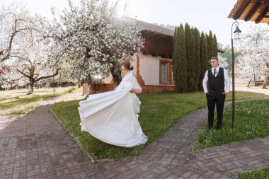 Gelin gelinliğinin içinde daireler çiziyor ve damat da ona bakıyor. Güzel bir bahar parkında düğün fotoğrafı çekimi..