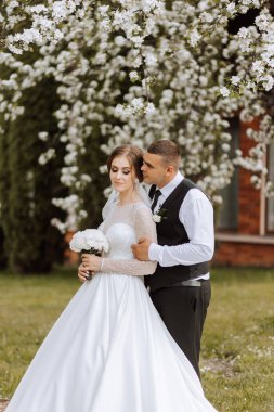 Düğün için. Aşk ve çift düğün için bahçede. Törenin ve bağlılığın kutlanması. Tarihi kaydet. Güven. Gelin ve damat kucaklaşır. Damat gelini bahar çiçekli bir ağacın altında kucaklıyor..