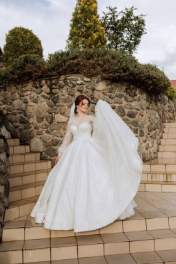 Gelin, muhteşem bir elbise ve duvak içinde, taş bir duvarın yanında poz veriyor. Uzun kollu muhteşem bir elbise, açık göğüs. Yaz düğünü