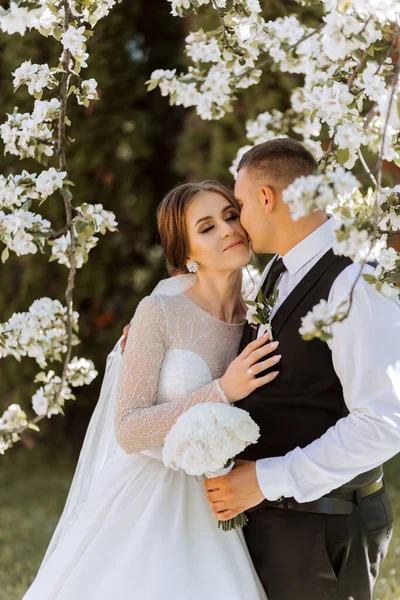 Düğün için. Aşk ve çift düğün için bahçede. Törenin ve bağlılığın kutlanması. Tarihi kaydet. Güven. Gelin ve damat kucaklaşır. Damat gelini bahar çiçekli bir ağacın altında kucaklıyor..