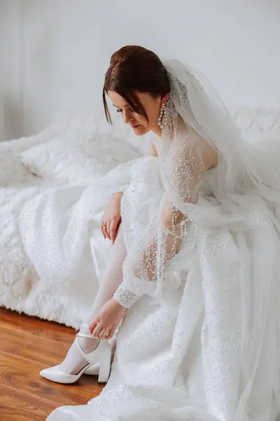 Young bride in beautiful wedding dress putting on shoes indoors, close-up. The bride in a wedding dress and a beautiful hairdo.
