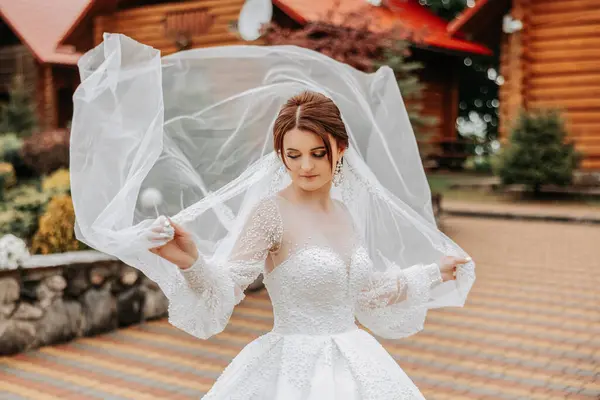A red-haired bride twirls her long veil against a background of wooden houses. Magnificent dress with long sleeves, open bust. Summer wedding