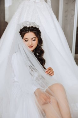 Mutlu bir gelin, bir otel odasında, bir mankenin üzerinde gelinliğiyle lüks düğününe hazırlanıyor. Şık saçlı, makyajlı ve sabahlık giymiş bir kadının portresi..