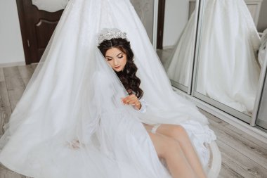 Mutlu bir gelin, bir otel odasında, bir mankenin üzerinde gelinliğiyle lüks düğününe hazırlanıyor. Şık saçlı, makyajlı ve sabahlık giymiş bir kadının portresi..