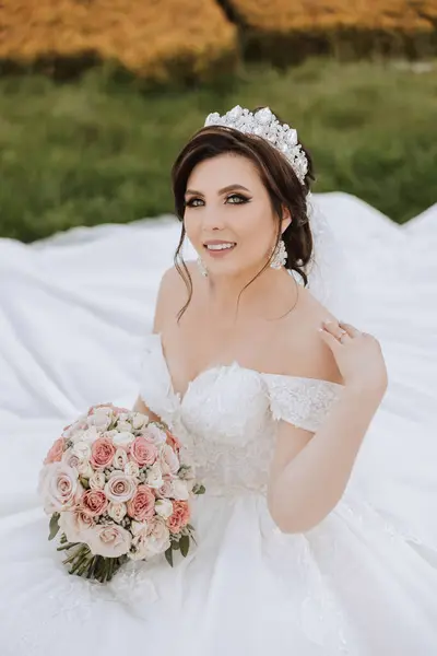 brunette bride in off-the-shoulder lace white dress and tiara posing with a bouquet of white and pink flowers while sitting on green grass. Beautiful hair and makeup. Spring wedding