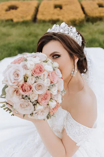 brunette bride in off-the-shoulder lace white dress and tiara posing with a bouquet of white and pink flowers. Beautiful hair and makeup. Spring wedding