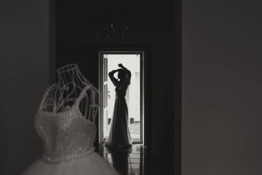 Mutlu bir gelin, bir otel odasında, bir mankenin üzerinde gelinliğiyle lüks düğününe hazırlanıyor. Şık saçlı, makyajlı ve sabahlık giymiş bir kadının portresi..