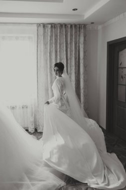 Güzel bir esmer gelin, güzel bir elbise içinde pencerenin kenarındaki gelinliğine doğru yürümeye hazırlanıyor. Siyah beyaz fotoğraf.