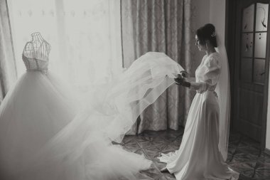 Güzel bir esmer gelin, pencerenin yanındaki gelinliğinin yanında güzel bir elbise içinde düğüne hazırlanıyor. Siyah beyaz fotoğraf.