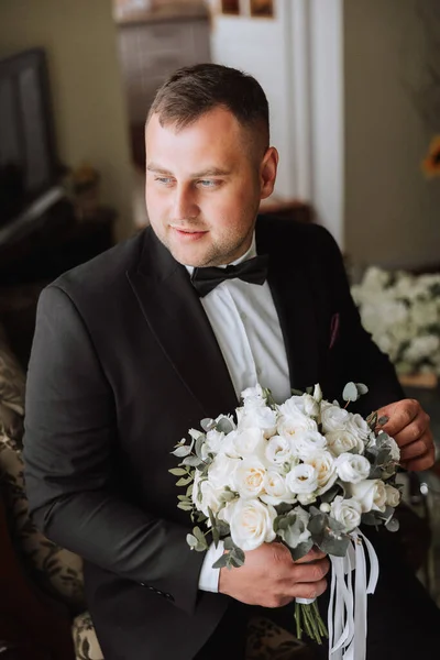 Stylish portrait of the groom with a bouquet of flowers. A man is preparing for a wedding ceremony in the morning. Groom's morning. Preparation for the groom's morning. Young and handsome groom.