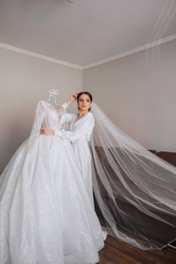 Mutlu bir gelin, bir otel odasında, bir mankenin üzerinde gelinliğiyle lüks düğününe hazırlanıyor. Şık saçlı, makyajlı ve sabahlık giymiş bir kadının portresi..
