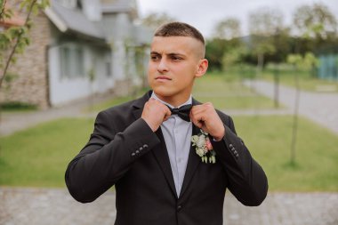 Siyah takım elbiseli damat papyonunu düzeltir, yeşil bir ağacın arka planına karşı poz verir. Düğün portresi.