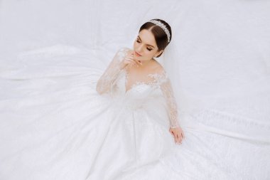 Uzun kollu dantelli beyaz elbiseli esmer bir gelin elbisesinin arka planına karşı duruyor. Beyaz bir arka planda. Klasik saç stili. Akşam makyajı. Bahar düğünü