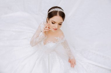 Uzun kollu dantelli beyaz elbiseli esmer bir gelin elbisesinin arka planına karşı duruyor. Beyaz bir arka planda. Klasik saç stili. Akşam makyajı. Bahar düğünü