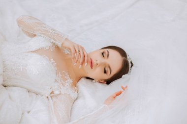 Uzun kollu dantelli beyaz elbiseli esmer bir gelin elbisesinin arka planına karşı duruyor. Beyaz bir arka planda. Klasik saç stili. Akşam makyajı. Bahar düğünü