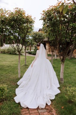 Uzun trenli dantelli beyaz elbiseli esmer gelin, bir buket beyaz ve pembe çiçekle ağaçların arkasına poz veriyor. Duvak havada. Güzel saç ve makyaj..