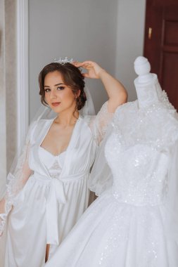 Mutlu bir gelin, bir otel odasında, bir mankenin üzerinde gelinliğiyle lüks düğününe hazırlanıyor. Şık saçlı, makyajlı ve sabahlık giymiş bir kadının portresi..