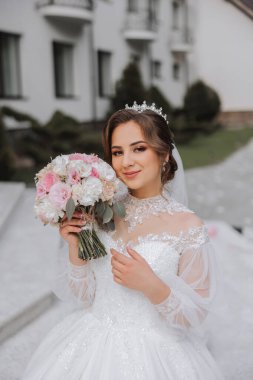 Omzundan ayrılmış esmer gelin dantelli beyaz elbise ve bir buket beyaz ve pembe çiçekle poz veren taç. Güzel saç ve makyaj. Bahar düğünü