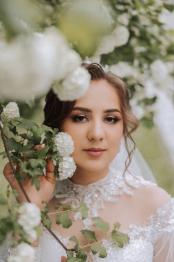 Kıvırcık esmer gelin, duvak ve uzun kollu elbisesiyle beyaz bir leylağın yanında poz veriyor. Güzel saç ve makyaj. Bahar düğünü