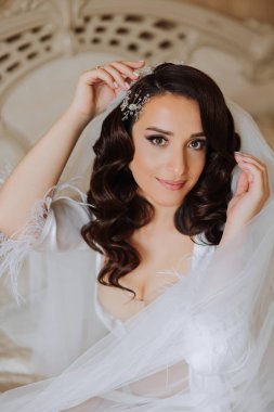 Saçında taç olan güzel bir esmer gelin, güzel bir bornozla düğüne hazırlanıyor. Gelinin yakın plan düğün portresi. Yatağa uzanmış, fotoğraf..