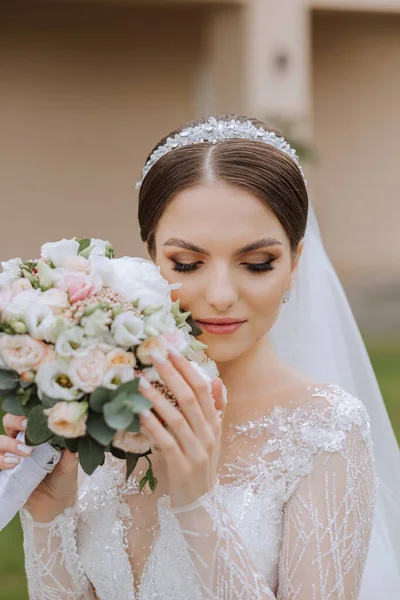 A brunette bride in a lacy white dress with long arms and a tiara poses with a bouquet of white and pink flowers. Beautiful hair and makeup. Spring wedding