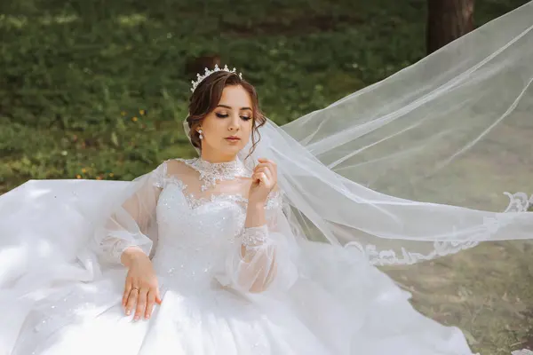 The bride in a lush dress with long sleeves poses with her veil in the air, posing while sitting on the grass, against a green background. Beautiful hairstyle, royal tiara. Spring wedding