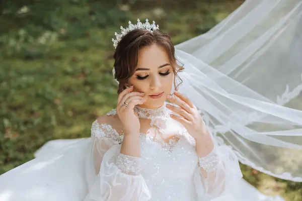 The bride in a lush dress with long sleeves poses with her veil in the air, posing while sitting on the grass, against a green background. Beautiful hairstyle, royal tiara. Spring wedding