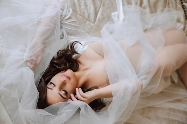 Stock image A beautiful brunette bride with a tiara in her hair is preparing for the wedding in a beautiful robe in boudoir style. Close-up wedding portrait of the bride lying on the bed, photo from above.