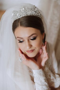 Beyaz dantelli gelinlik içinde güzel, genç, mükemmel bir gelin sabah yatak odasında oturuyor ve kulağına küpe takıyor. Düğün fotoğrafı, portre, konsept.