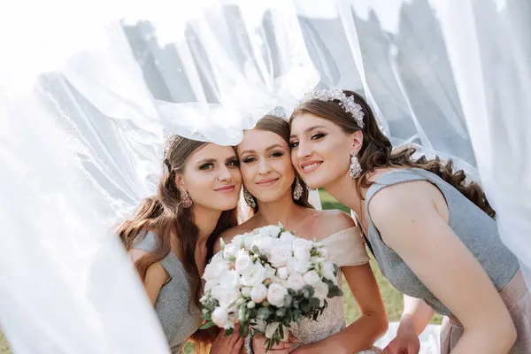 Bridesmaids are wrapped in a veil. The bride is holding a bouquet. Long veil. Wedding in nature. Smiling girls in identical dresses.