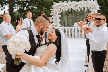 Gelin ve damat, arkadaşları tören kemerinin yanında. Arkadaşlar gelinle damadı destekliyor. Gösterişli ve zarif bir gelin elbisesi. Şık bir düğün. Doğada yaz düğünü. Damat gelini öper.