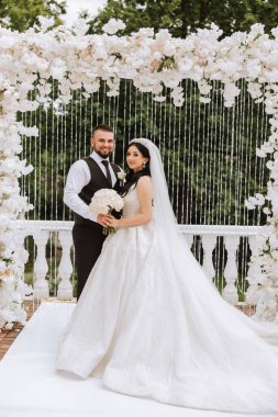 Yakışıklı bir damat, gelinini bembeyaz bir elbiseyle kucaklıyor ve güzel bir açık havada gülümsüyor. Açık gökyüzünün altında. Yüksek kalite fotoğraf. Yeni evli bir çift güneşli bir yaz gününde birlikte poz verir..