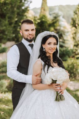 Yakışıklı bir damat, gelinini bembeyaz bir elbiseyle kucaklıyor ve güzel bir açık havada gülümsüyor. Açık gökyüzünün altında. Yüksek kalite fotoğraf. Yeni evli bir çift güneşli bir yaz gününde birlikte poz verir..