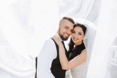 Yakışıklı bir damat, gelinini bembeyaz bir elbiseyle kucaklıyor ve güzel bir açık havada gülümsüyor. Açık gökyüzünün altında. Yüksek kalite fotoğraf. Yeni evli bir çift güneşli bir yaz gününde birlikte poz verir..