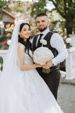 Yakışıklı bir damat, gelinini bembeyaz bir elbiseyle kucaklıyor ve güzel bir açık havada gülümsüyor. Açık gökyüzünün altında. Yüksek kalite fotoğraf. Yeni evli bir çift güneşli bir yaz gününde birlikte poz verir..