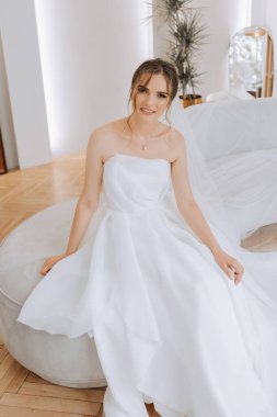 Otel odasında gelinin portresi. Güzel genç bir kız beyaz bir gelinlik giyiyor. Modern düğün saç stili. Doğal makyaj..