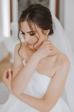 Otel odasında gelinin portresi. Güzel genç bir kız beyaz bir gelinlik giyiyor. Modern düğün saç stili. Doğal makyaj..