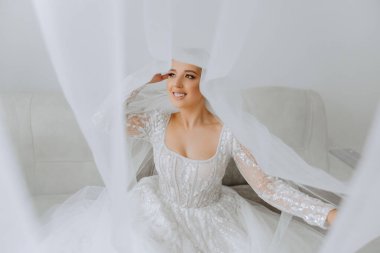 Otel odasında gelinin portresi. Güzel genç bir kız beyaz bir gelinlik giyiyor. Modern düğün saç stili. Doğal makyaj..