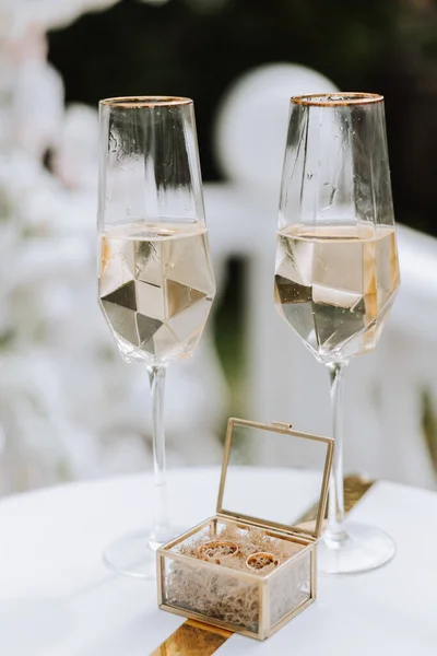 Shaped champagne glasses and a glass box with gold wedding rings in the middle. Preparation for the wedding ceremony.