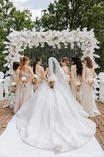 The bride and her bridesmaids turned their backs to the camera against the background of green trees. A long train on the dress. The girls are wearing identical dresses at the wedding.
