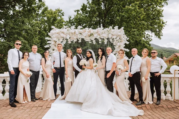 The bride and groom and their friends pose near the arch. Long train of the dress. Stylish wedding. Summer wedding in nature