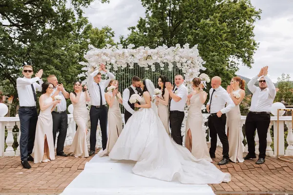 The bride and groom and their friends pose near the arch. Long train of the dress. Stylish wedding. Summer wedding in nature