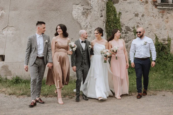 The bride and groom with their friends pose on the old wall. A beautiful and elegant dress of the bride. Stylish wedding. Summer wedding in nature